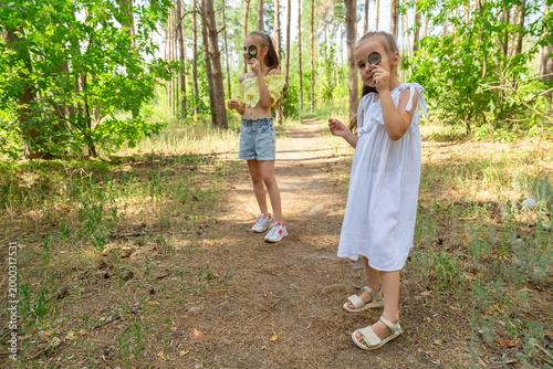 Two kids in casual clothes in summer exploring nature in forest together. Little girl looking through magnifying glass while squatting on ground