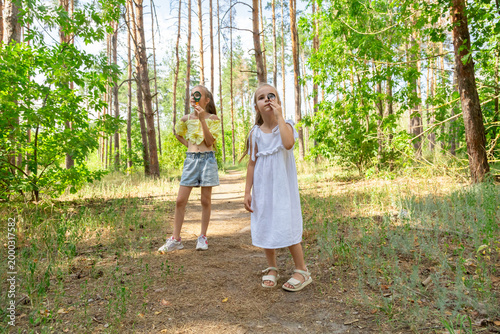 Two kids in casual clothes in summer exploring nature in forest together. Little girl looking through magnifying glass while squatting on ground