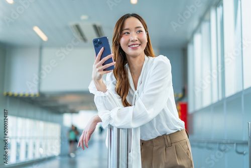 Asian Businesswoman leaning on railing, holding up smartphone in airport walkway