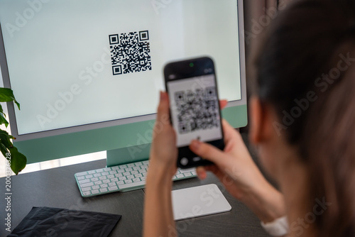 Person scanning QR code from computer screen with smartphone at home workspace. Digital interaction between devices and online service access concept.