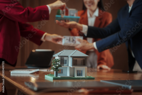 фотографія Close-up of hands exchanging keys and money over a table with model house, real estate purchase agreement meeting, agents and buyers in background, warm indoor office
