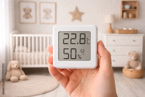 Hand holds a digital thermometer hygrometer in a baby room to check indoor comfort, warmth, and moisture levels.