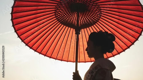 A silhouette of a woman with traditional Japanese hairdo holding a large, red paper parasol against a sunset sky