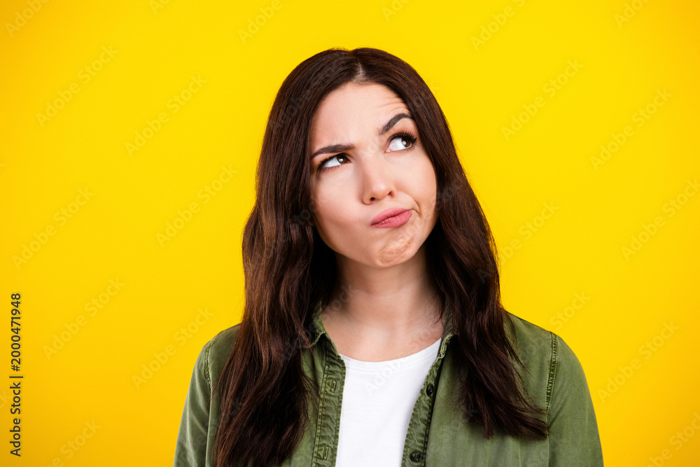 Fototapeta premium Pensive young woman looking up with a puzzled expression against a bright yellow background, portrait conveying doubt, thinking and indecision in casual green shirt