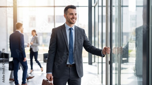 Smiling businessman entering office building with briefcase colleagues in background modern interior