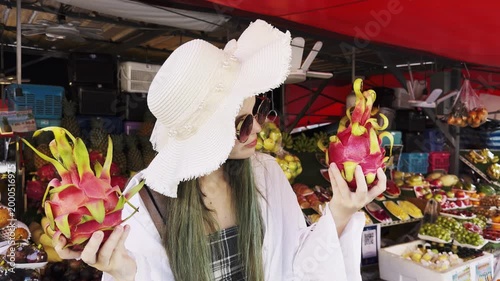 Young woman in sun hat holding two dragon fruits at tropical market. Fresh exotic fruits, summer lifestyle, travel vibes and healthy nutrition concept in vibrant street market scene