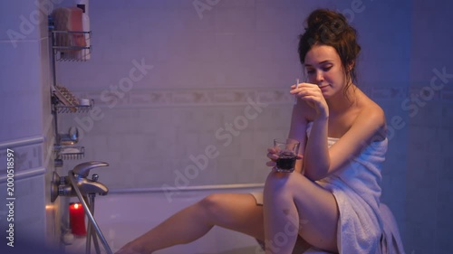Melancholic young woman wrapped in a towel sits on the edge of a bathtub, holding a glass of alcohol and looking distressed at home