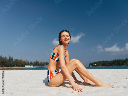 Smiling young woman sits on white sand at the beach, wearing a colorful striped bikini under a clear blue sky. Sunlit, relaxed pose, warm mood, casual outdoor portrait.