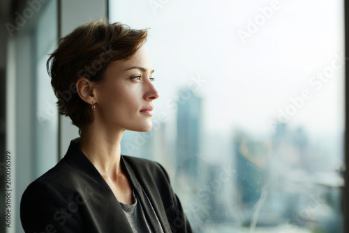 Confident businesswoman standing near office window looking forward, city skyline softly blurred in background, natural daylight illumination, professional corporate lifestyle scene, visual for social