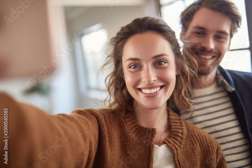 Clean background featuring smiling woman taking selfie with partner standing close beside her, cozy home interior softly blurred, warm natural window light, authentic lifestyle moment, in clean