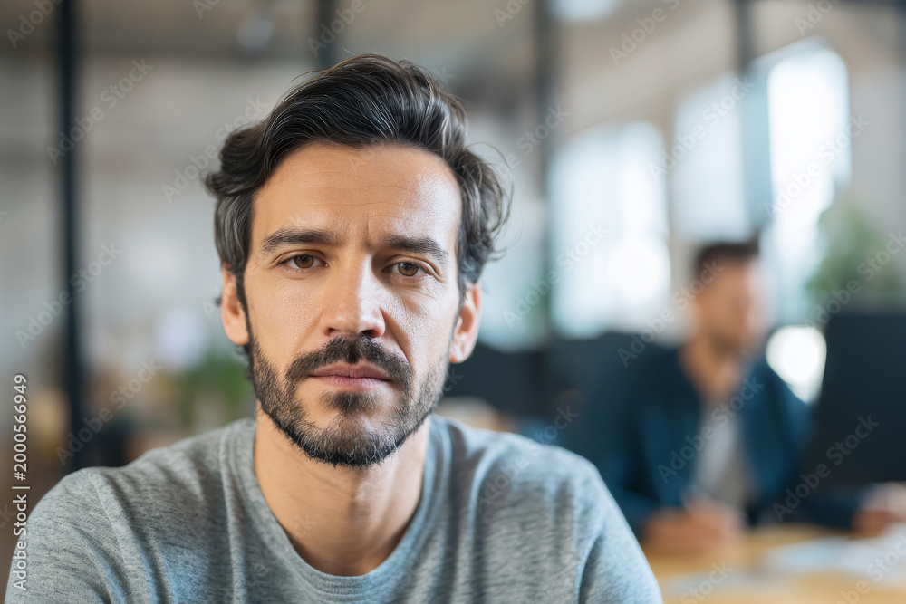 Obraz premium Detailed view of man looking at camera with calm confident expression, modern workspace background softly blurred with coworker present, natural daylight illumination, with blank space
