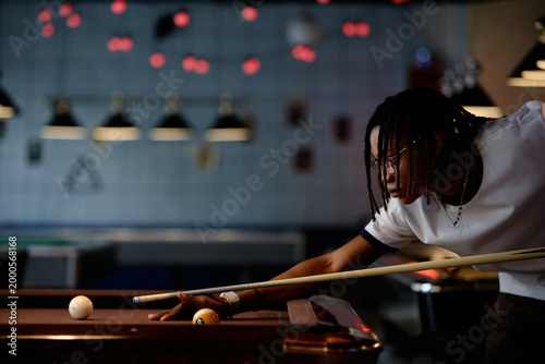 Black young adult man aiming pool cue over billiards table, focusing on precision shot during leisure game. Ideal for recreation marketing, nightlife promotion, competitive hobby content
