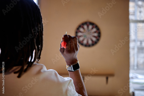 Black young adult woman aiming dart at board, practicing focus and leisure game indoors. Useful for recreation marketing, concentration training, lifestyle wellness campaigns
