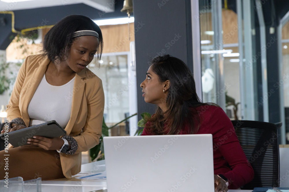 Fototapeta premium Diverse female team in tan blazer burgundy top leaning with tablet discussing office laptop papers