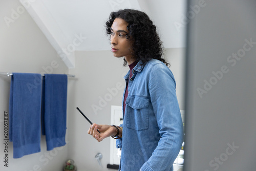 Nonbinary adult in denim shirt with bracelets holding eyeliner, looking at mirror at bathroom sink