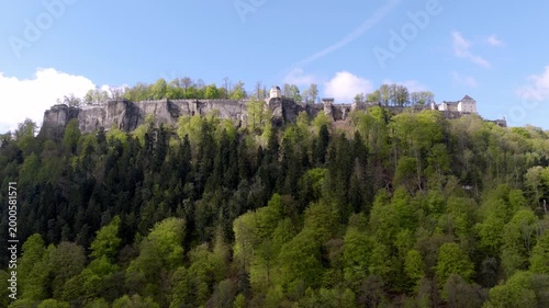 Aerial view of Königstein Fortress in Saxon Switzerland atop a steep sandstone rock, surrounded by dense spring forest under a blue sky.