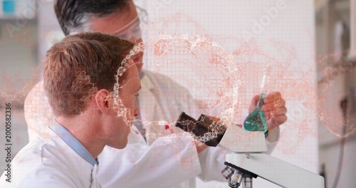 Leaning scientist peering into microscope in lab, mentor holding green flask with overlay, in coats