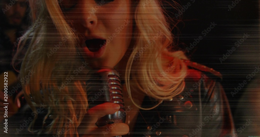 Fototapeta premium Singing female singer holding retro mic on club stage, wearing studded jacket, guitar neck visible