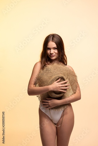 Young woman posing in swimwear with a large straw hat, representing summer fashion, confidence, and trendy beach style