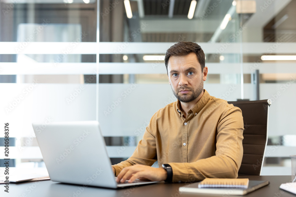 Fototapeta premium Man focusing on his laptop in a modern corporate office, looking directly at the camera with a serious expression while engaged in daily professional tasks and online communication