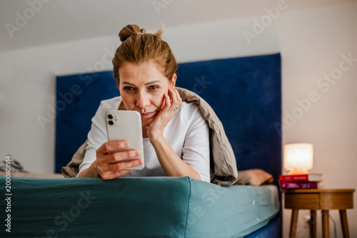 Fotografija Woman lying under blanket and scrolling on phone.