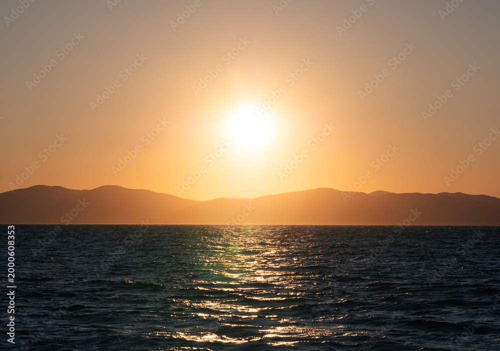 Naklejka premium Sun dips below distant mountains painting the vast ocean with a warm, fiery glow at dusk