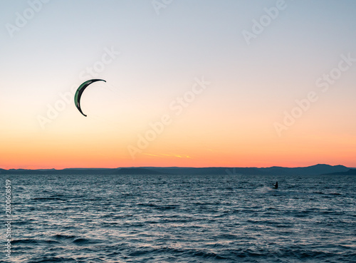 Kitesurfer rides ocean waves with a vibrant kite under a beautiful gradient sunset sky and distant mountains