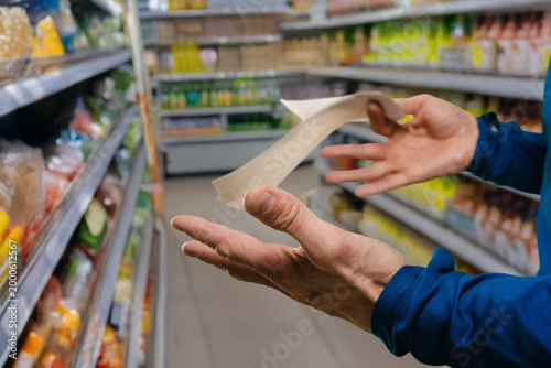Bald caucasian man holding long supermarket receipt with visible surprise near stocked shelves, closeup of hands and paper roll, blue top contrasted with aisle products, reacting