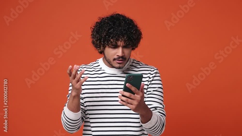 Sad upset young Indian man wear striped casual clothes hold use mobile cell phone swear read bad fake news unexpected rumor has some problem isolated on plain orange red background. Lifestyle concept