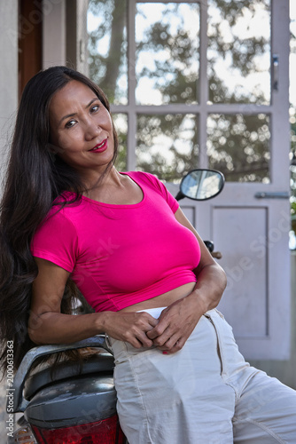 Asian girl in pink top sitting on ledge outdoors legs crossed looking away thoughtfully