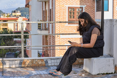 Woman in black outfit sitting on rooftop ledge using smartphone urban skyline backdrop