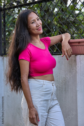Young woman in pink top and white jeans leaning against wall outdoors looking off-camera thoughtfully