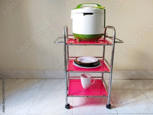 A three-tier stainless steel kitchen serving trolley with a white and green rice cooker on top, a stack of plates in the middle and porcelain tea cups at the bottom.