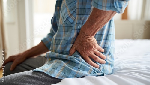 An elderly man experiencing stomach pain, clutching his abdomen while sitting on a bed in a bright room, conveying a sense of discomfort and distress