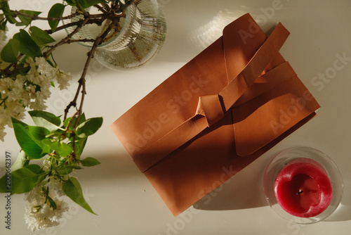 Gift box with copper wrapping and ribbon beside a candle and greenery in a cozy home setting, offering a warm still life for festive decor, craft and interior design.