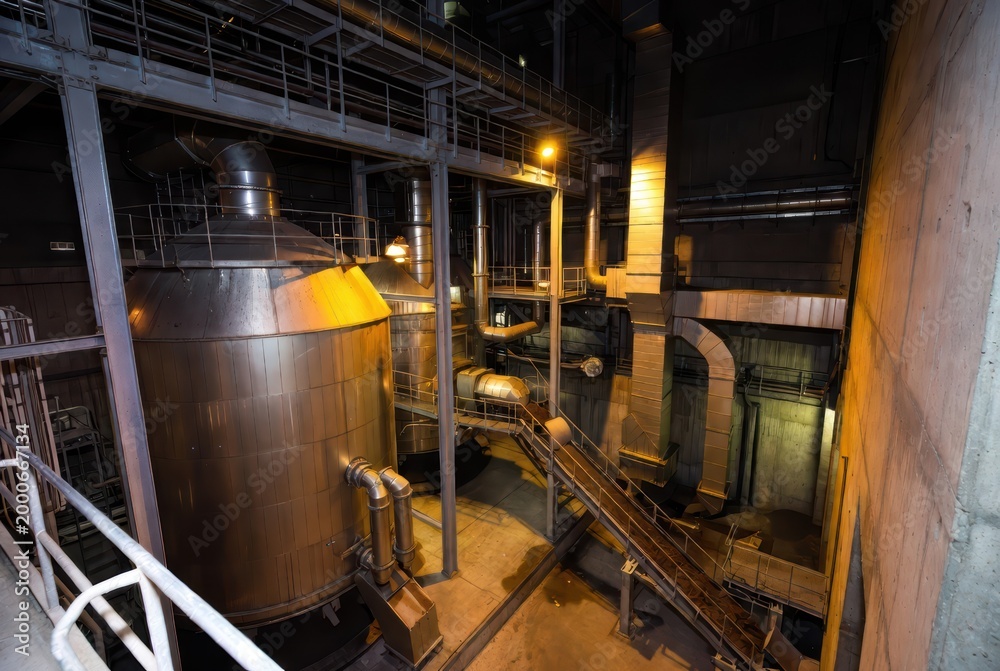 Fototapeta premium Industrial interior with large metal tanks, pipes, and walkways, illuminated by warm lights.