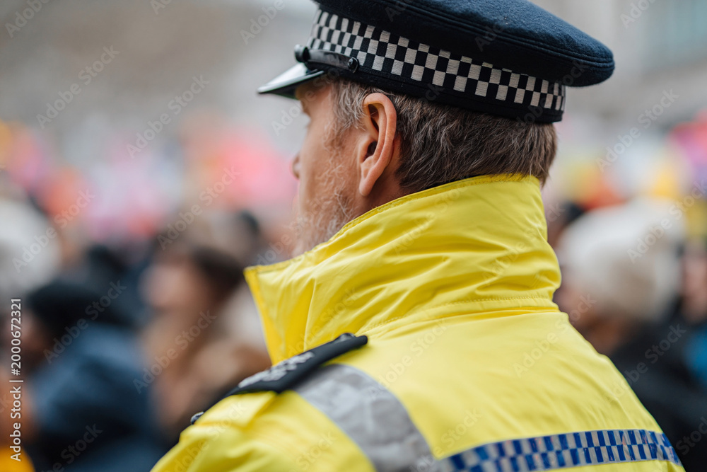 Fototapeta premium Yellow jacket, checkered cap. Back view, focused on shoulder gear. Crowd blurred, suggests public event. Neutral expression, duty-focused gaze. Ideal for safety, authority, civic scene