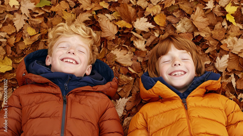 Two cheerful boys lie on a pile of autumn leaves, smiling happily at the camera