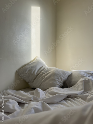 Soft morning sunlight illuminating cozy unmade bed with white pillows and wrinkled linen sheets creating calm minimal bedroom interior atmosphere