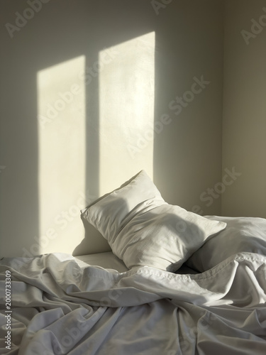 Warm sunlight casting window shadows on unmade bed with soft white pillows and wrinkled linen sheets creating peaceful minimalist bedroom interior scene