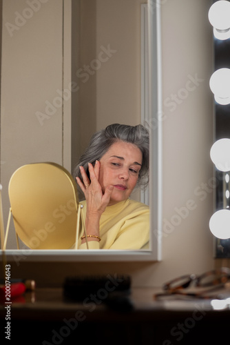 Senior woman with gray hair examines her face in a bright dressing-room mirror, practicing a gentle skincare routine and self-care ritual in a calm, domestic setting