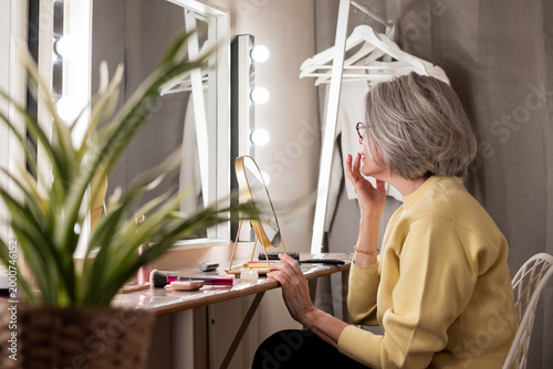 Senior woman sitting at a modern vanity table, applying lipstick while looking into the mirror with bright lights, focusing on her morning routine and self-care
