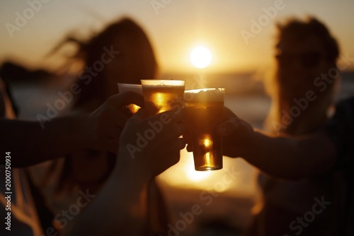 People raise drinks to celebrate sunset light at the beach near the ocean in warm weather