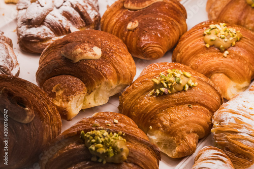 Fotografie Shallow-depth-of-field close-up of a bakery display featuring multiple golden-brown croissants and cornetti topped with crushed green pistachios, cream filling, and powdered sugar