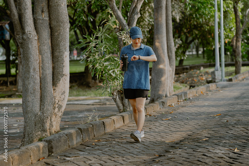 Asian women jogging in park holding a water bottle drink while checking smartwatch, representing fitness tracking, active lifestyle and health monitoring during outdoor cardio workout.