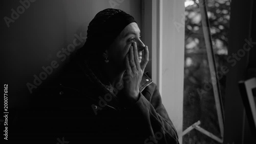 Woman undergoing cancer treatment wipes tears by window, black and white scene capturing grief, emotional pain and vulnerability during a quiet moment at home
