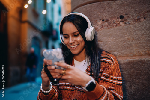 Happy multiracial girl texting on mobile phone wearing headphones, young hispanic female using smartphone audio app in urban setting, lady checking social media while listening to podcast.