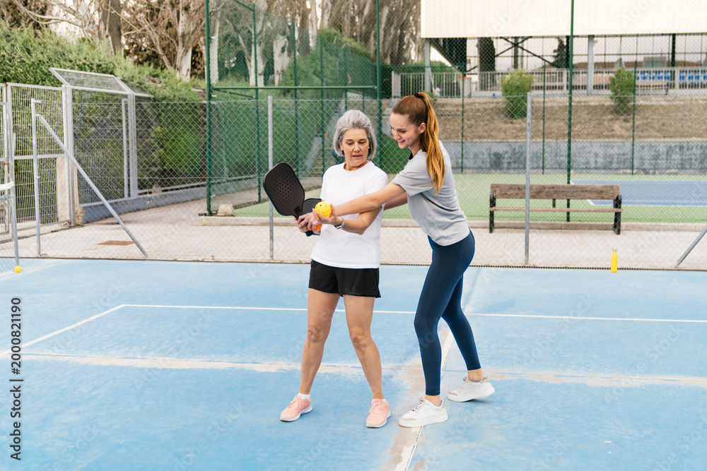 Obraz premium Young female coach guiding an older woman on playing pickleball, showing paddle grip and ball placement on a blue court