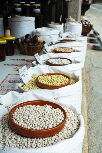 Traditional outdoor market display featuring rows of white sacks filled with various dried beans and legumes. Clay bowls and price tags highlight a rustic, organic grocery shopping experience