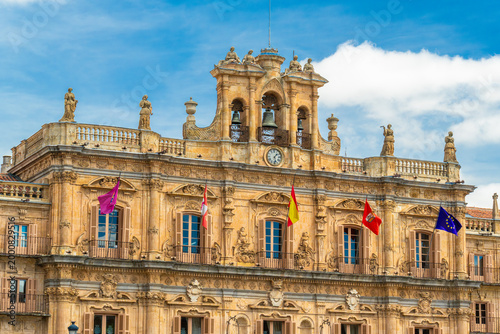 The ornate Baroque facade of the Salamanca City Hall (Ayuntamiento) in Plaza Mayor, Spain. Features intricate stone carvings, iron balconies, statues, and colorful flags under a blue sky